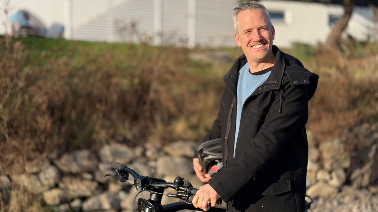 Mann står og holder en sykkel mens han smiler mot kamera. 