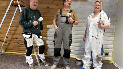 To menn og en kvinne i arbeidstøy som står foran en vegg som er malt halveis i hvitt. De tre personene holder opp hver sin malekost. Munter stemning. 