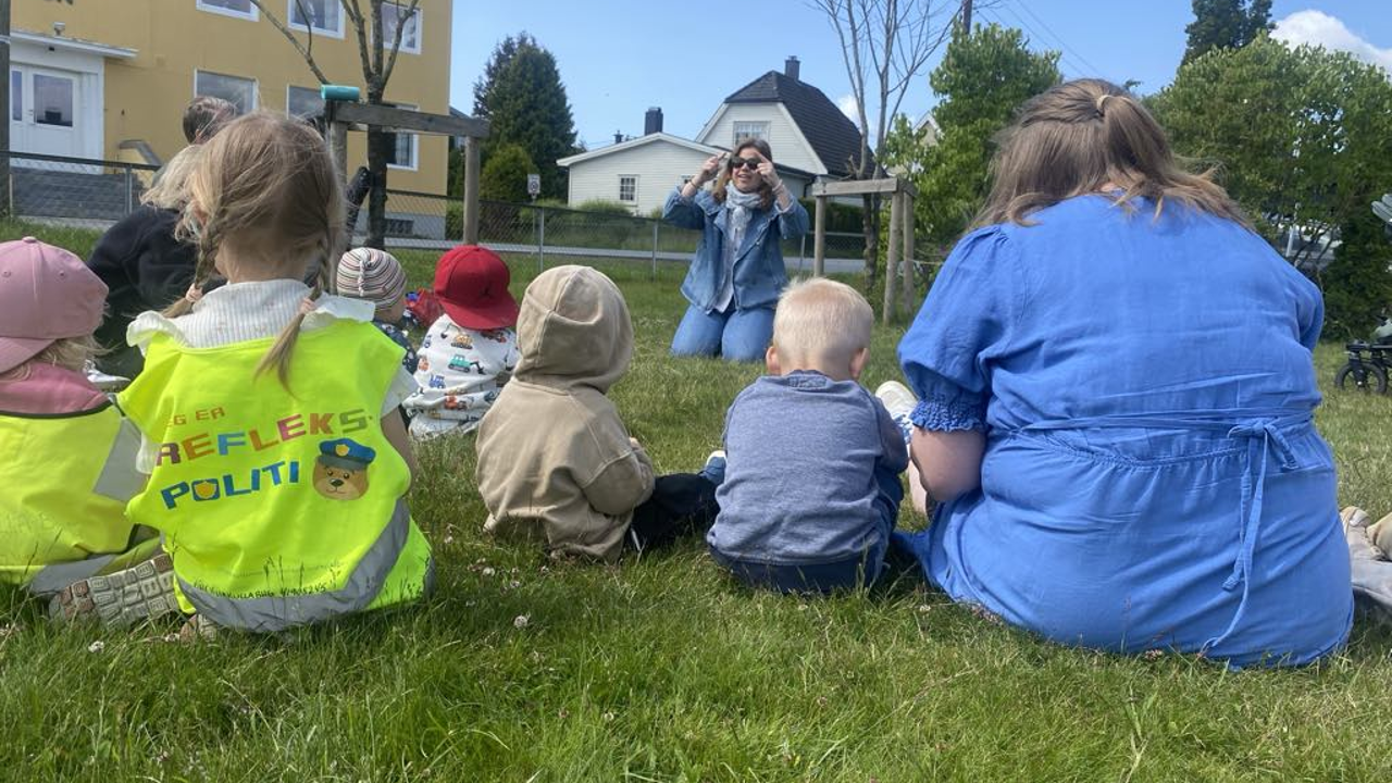Barn sitter på gresset og følger med på en voksen som leder musikksamling.
