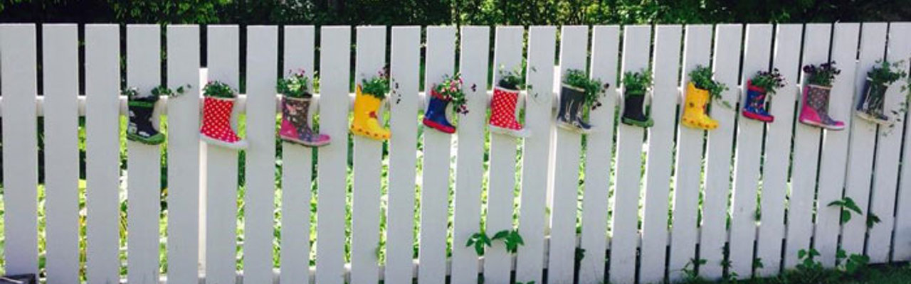 Fargerike gummistøvler hengende fra et hvitt gjerde. Gummistøvlene har fargerike blomster i seg, som en gummistøvel-vase.